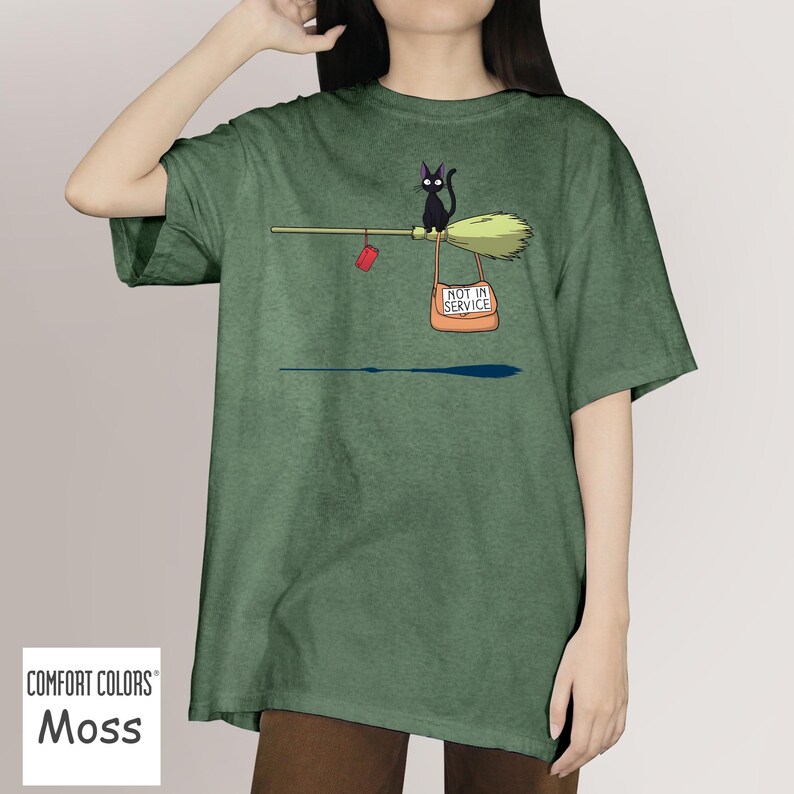 May include: Moss green Comfort Colors t-shirt featuring a black cat riding a broom with a red mail pouch and a sign that reads "NOT IN SERVICE". The shirt has a relaxed fit.