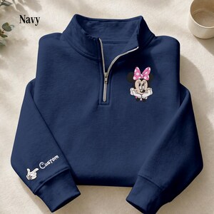 Pode incluir: Camisola azul marinho com fecho de correr de um quarto com um gr&aacute;fico da Minnie Mouse com um la&ccedil;o rosa com bolinhas. A manga esquerda tem a palavra "Custom" e um gesto de m&atilde;o. Z&iacute;per prateado.