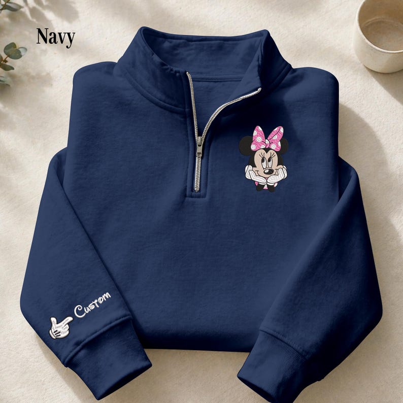 Pode incluir: Camisola azul marinho com fecho de correr de um quarto com um gr&aacute;fico da Minnie Mouse com um la&ccedil;o rosa com bolinhas. A manga esquerda tem a palavra "Custom" e um gesto de m&atilde;o. Z&iacute;per prateado.
