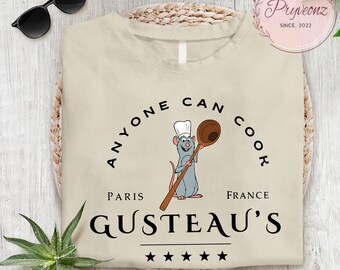 May include: Beige t-shirt featuring a cartoon rat chef holding a wooden spoon. The text "ANYONE CAN COOK" arches above the rat, with "PARIS FRANCE" and "GUSTEAU'S" below. Black sunglasses rest on the top left.