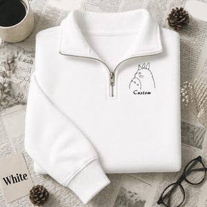 Könnte beinhalten: Weißes Sweatshirt mit Viertelreißverschluss, schwarzer Umrissillustration einer Zeichentrickfigur und dem Wort "Custom" darunter. Das Sweatshirt hat einen Kragen und gerippte Bündchen. Das Wort "White" ist auf einer kleinen Karte aufgedruckt.