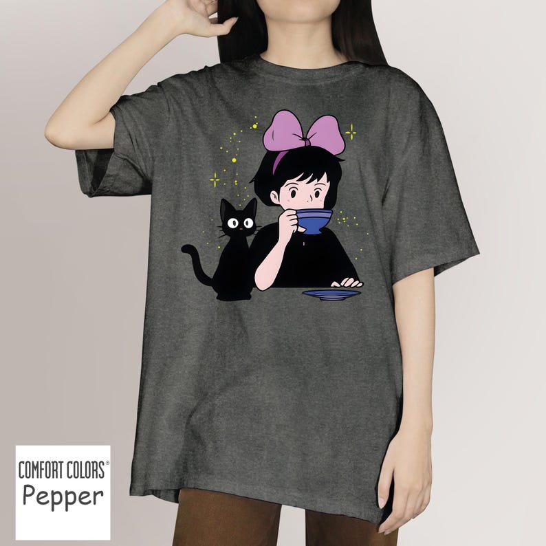 May include: Dark gray Comfort Colors t-shirt featuring a cartoon illustration of a girl with a pink bow, drinking from a blue cup, with a black cat. The design includes yellow star accents.