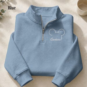 May include: A light blue pullover sweatshirt with a quarter-zip neckline. The sweatshirt features a white outline of a mouse head and the word "Custom" in script. The word "Blue" is printed in the upper left corner.