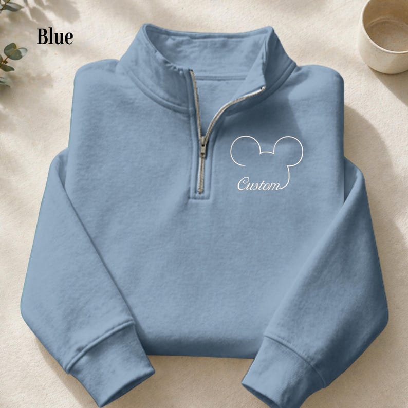 May include: A light blue pullover sweatshirt with a quarter-zip neckline. The sweatshirt features a white outline of a mouse head and the word "Custom" in script. The word "Blue" is printed in the upper left corner.