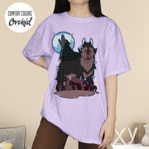 May include: A light purple t-shirt with a graphic of a wolf pack under a full moon. The design features a large wolf with red eyes and a smaller wolf howling at the moon. The shirt has the words "COMFORT COLORS" and "Orchid" in a circle.
