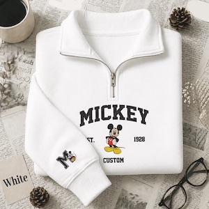 May include: A white quarter-zip sweatshirt featuring a black collegiate-style MICKEY text graphic, a Mickey Mouse illustration, the text EST. 1928, and the word CUSTOM. The sleeve displays a large letter M with a small Mickey Mouse graphic.