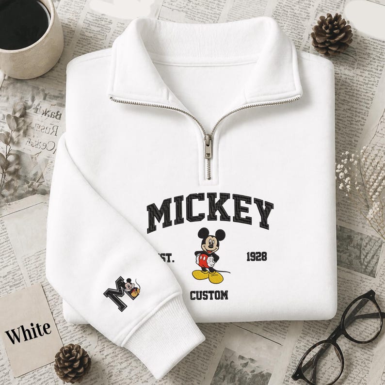 May include: A white quarter-zip sweatshirt featuring a black collegiate-style MICKEY text graphic, a Mickey Mouse illustration, the text EST. 1928, and the word CUSTOM. The sleeve displays a large letter M with a small Mickey Mouse graphic.