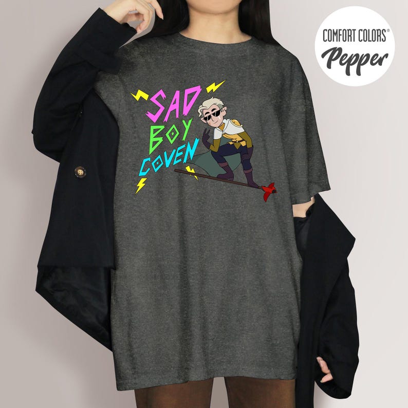 Puede incluir: Camiseta Comfort Colors gris oscuro con el texto "SAD BOY COVEN" en rosa, verde y azul, con rayos amarillos. La camiseta presenta una ilustraci&oacute;n de dibujos animados de un personaje con gafas de sol.