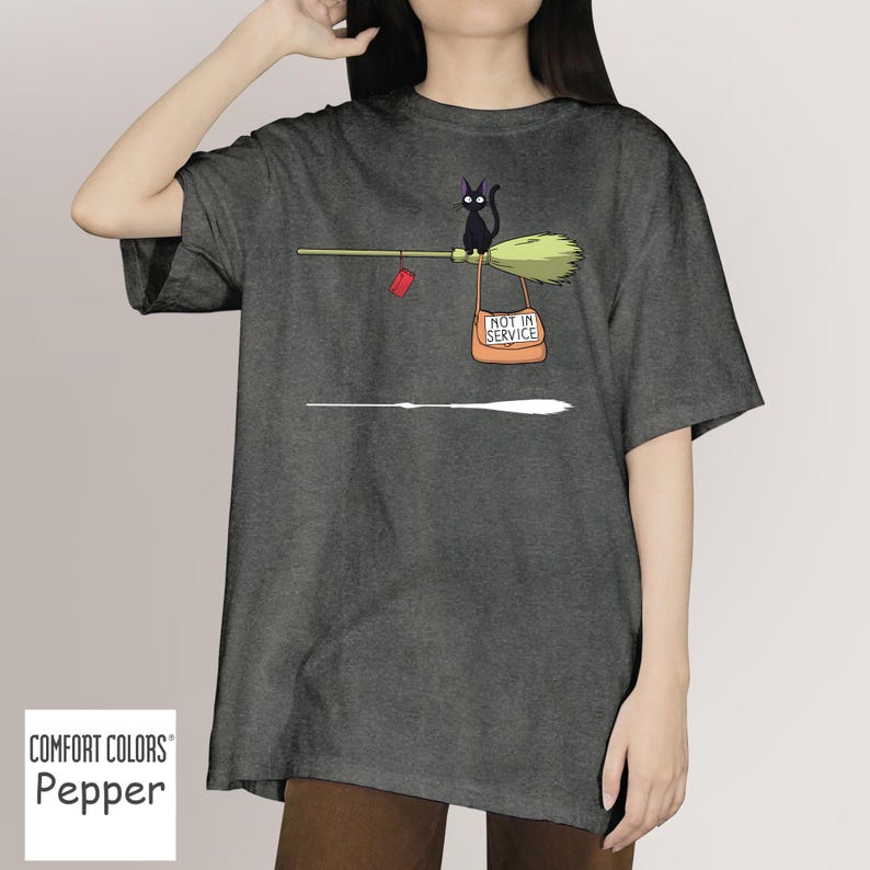 May include: Dark grey t-shirt with a whimsical graphic. A black cat sits on a broom with a sign that reads "NOT IN SERVICE." A red package hangs from the broom. The shirt is labeled "Comfort Colors Pepper."