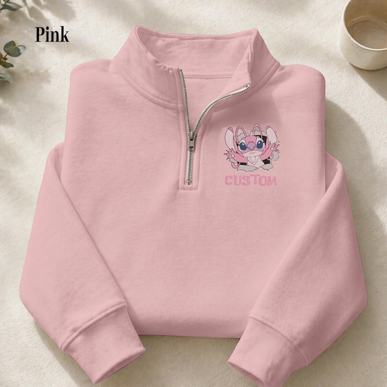 May include: A light pink quarter-zip sweatshirt with a cartoon character graphic and the word "CUSTOM" printed on the front. The sweatshirt has a silver zipper and long sleeves. The word "Pink" is in the upper left corner.
