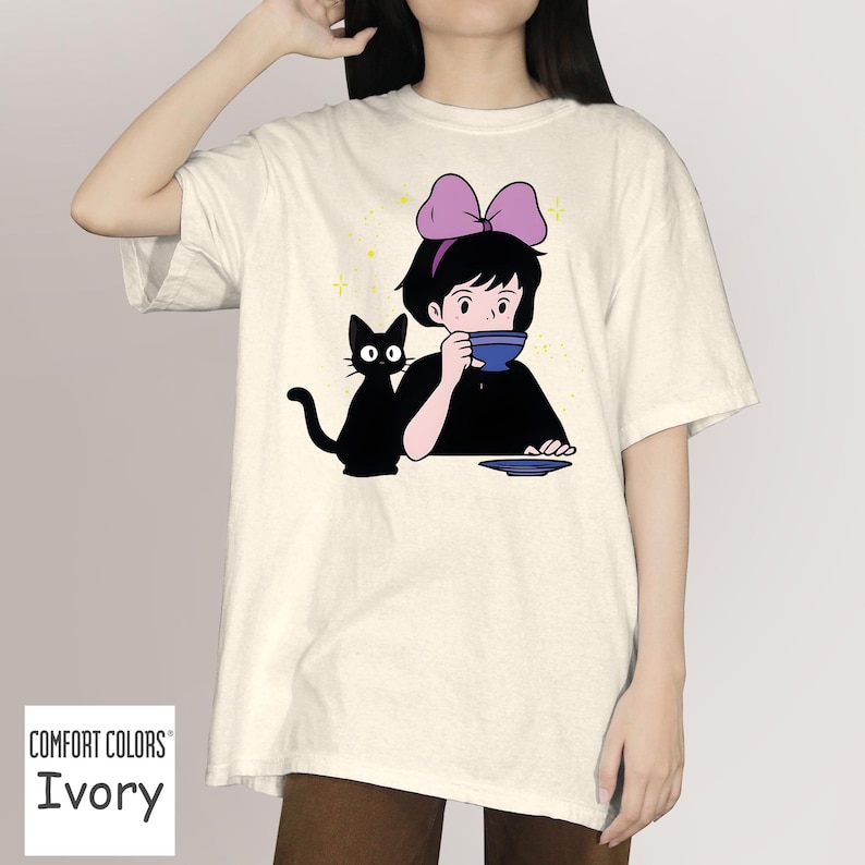 May include: Ivory Comfort Colors t-shirt featuring a cartoon illustration of a girl with a purple bow drinking from a blue cup, a black cat sits beside her. The shirt has the word "Ivory" printed at the bottom.