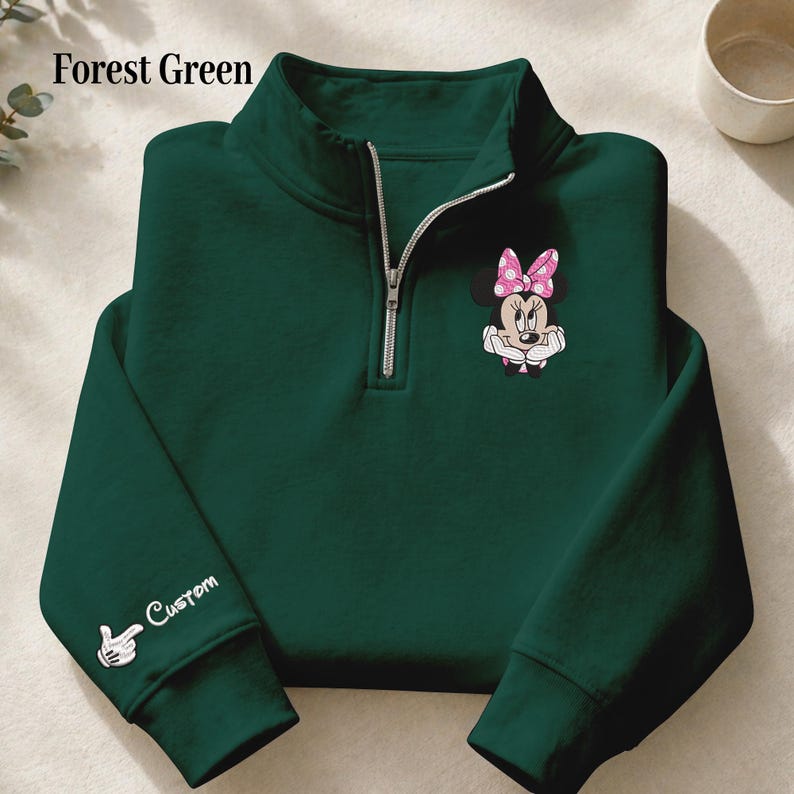 Pode incluir: Camisola verde floresta com fecho de correr de um quarto com um gr&aacute;fico da Minnie Mouse com um la&ccedil;o rosa de bolinhas. A manga esquerda tem um design personalizado com a palavra "Custom" e uma m&atilde;o com um dedo a apontar. Fecho de correr prateado.