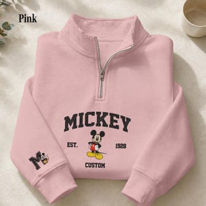 May include: A light pink quarter-zip sweatshirt featuring a black embroidered Mickey Mouse graphic on the chest with the text MICKEY, EST. 1928, and CUSTOM. The right sleeve displays a black letter M with a small Mickey Mouse icon inside the letter.