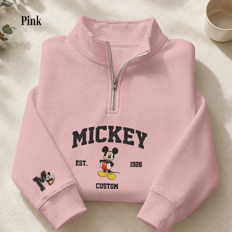 May include: A light pink quarter-zip sweatshirt featuring a black embroidered Mickey Mouse graphic on the chest with the text MICKEY, EST. 1928, and CUSTOM. The right sleeve displays a black letter M with a small Mickey Mouse icon inside the letter.