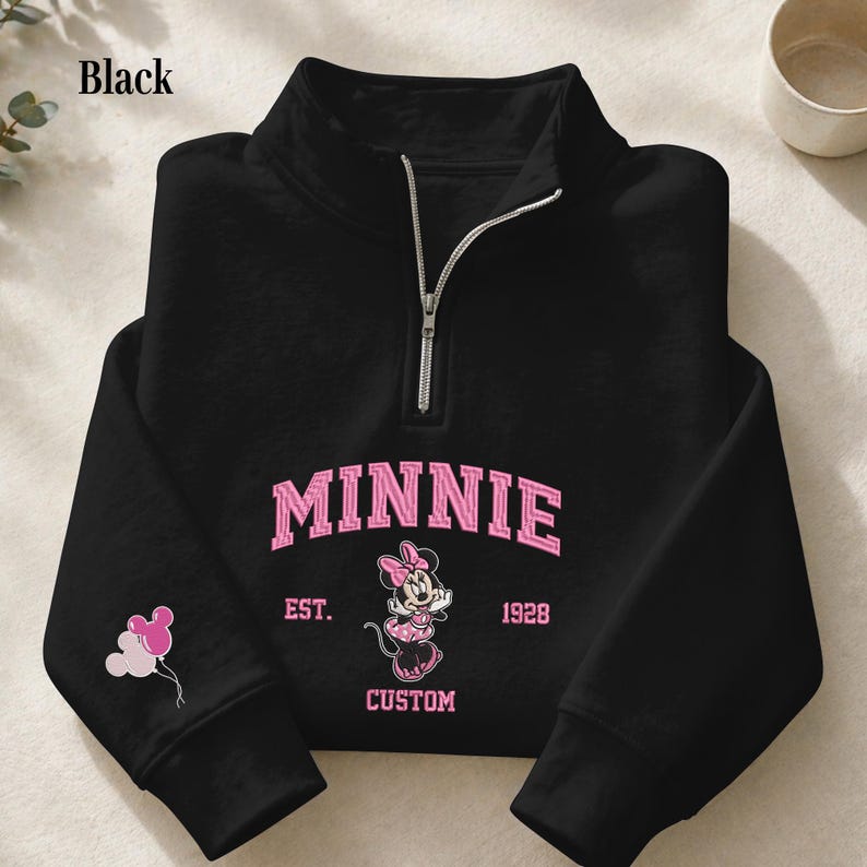 May include: A black quarter-zip sweatshirt featuring a pink graphic of Minnie Mouse with the text MINNIE EST. 1928 CUSTOM. The sleeve displays a small graphic of three balloons shaped like mouse ears in shades of pink and white.
