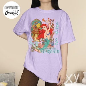May include: A light purple t-shirt featuring a colorful graphic of Ariel from The Little Mermaid with Flounder. The shirt has the words "The Little Mermaid" printed on it. The shirt also has the words "Comfort Colors" and "Orchid" printed on it.