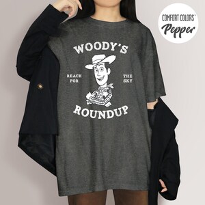 Pu&ograve; includere: T-shirt grigio scuro con una grafica di Woody di Toy Story, con la scritta "Woody's Roundup" e "Reach for the Sky". La maglietta &egrave; una maglietta Comfort Colors Pepper.