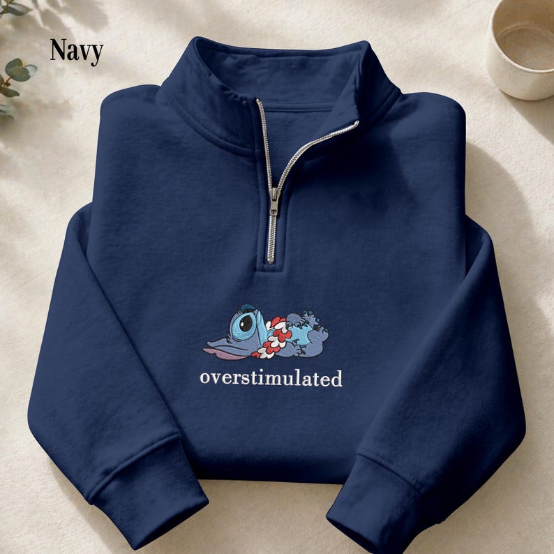 May include: A navy blue quarter-zip sweatshirt featuring a graphic of a blue alien character wearing a red and white floral lei, lying down with the text "overstimulated" printed in white lettering underneath the character on the chest.