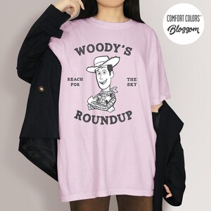 Pu&ograve; includere: T-shirt rosa chiaro con una grafica in bianco e nero di Woody di Toy Story, con il testo "WOODY'S ROUNDUP" e "REACH FOR THE SKY". La maglietta ha anche i loghi "Comfort Colors" e "Blossom".