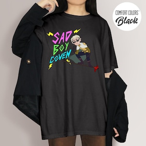 Puede incluir: Camiseta negra con el texto "SAD BOY COVEN" en rosa, verde y azul, con rayos amarillos. La camiseta presenta una ilustraci&oacute;n de dibujos animados de un personaje con gafas de sol y un atuendo amarillo y marr&oacute;n.