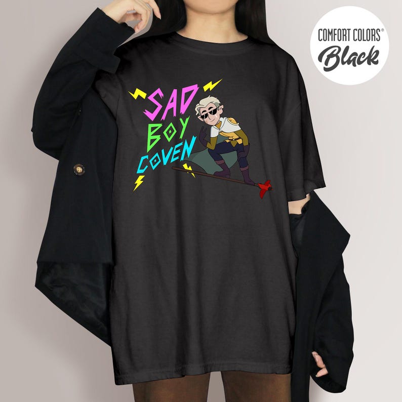 Puede incluir: Camiseta negra con el texto "SAD BOY COVEN" en rosa, verde y azul, con rayos amarillos. La camiseta presenta una ilustraci&oacute;n de dibujos animados de un personaje con gafas de sol y un atuendo amarillo y marr&oacute;n.