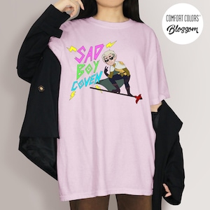 Puede incluir: Camiseta rosa claro con el texto "SAD BOY COVEN" en amarillo y verde, y una ilustraci&oacute;n de dibujos animados de un personaje con gafas de sol. La camiseta es de Comfort Colors y tiene un logotipo redondo.