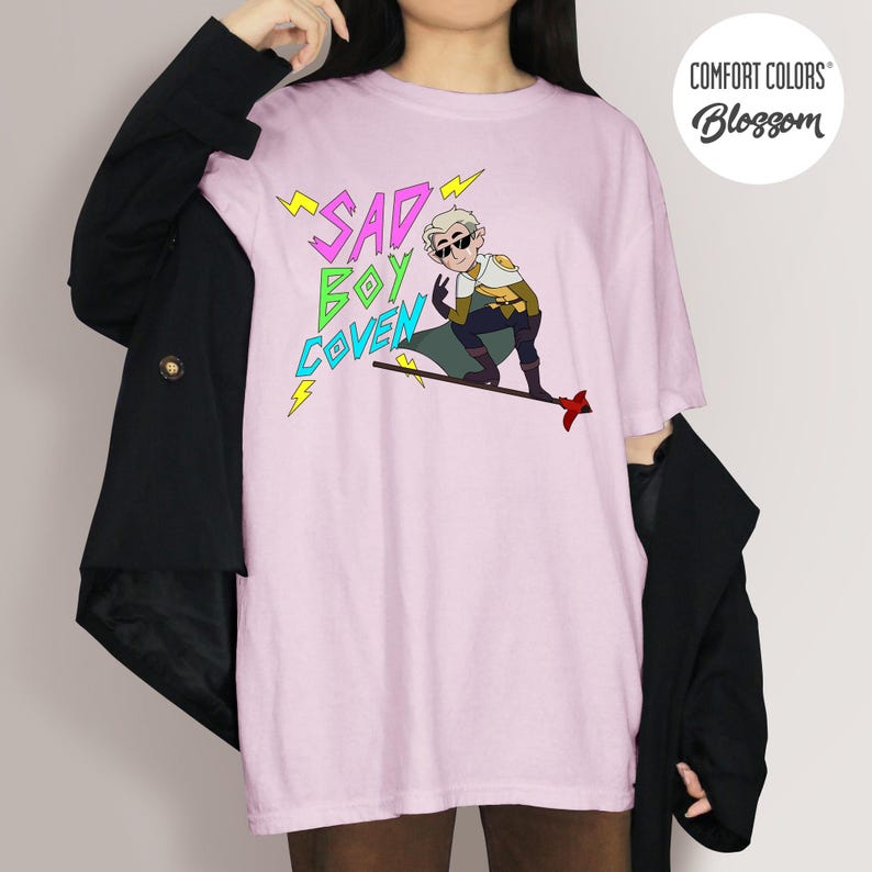 Puede incluir: Camiseta rosa claro con el texto "SAD BOY COVEN" en amarillo y verde, y una ilustraci&oacute;n de dibujos animados de un personaje con gafas de sol. La camiseta es de Comfort Colors y tiene un logotipo redondo.