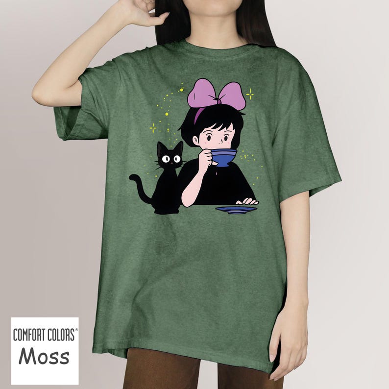 May include: Moss green Comfort Colors t-shirt featuring a cartoon illustration of a girl drinking from a blue cup, with a black cat beside her. The girl wears a pink bow. The shirt has a relaxed fit.