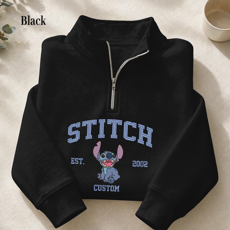 Pu&ograve; includere: Felpa nera con collo a zip a un quarto. La parte anteriore presenta la scritta "STITCH" in azzurro, un'immagine del personaggio Stitch e il testo "EST. 2002 CUSTOM". La felpa ha maniche lunghe.