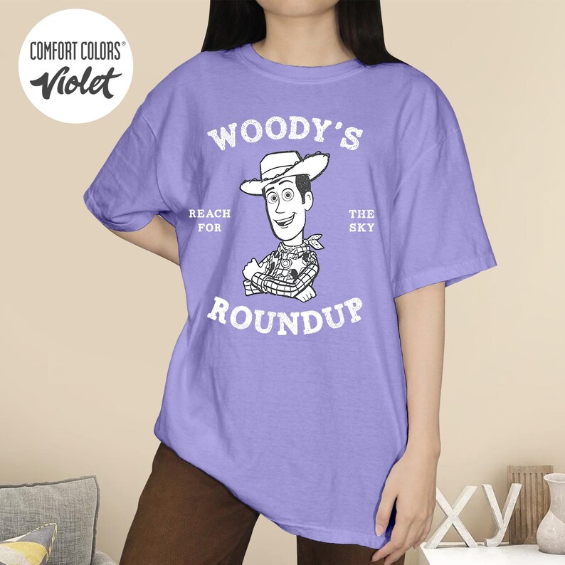 Pu&ograve; includere: T-shirt Comfort Colors viola con grafica di Woody di Toy Story. Il design include il testo "Woody's Roundup" e "Reach for the Sky". La maglietta &egrave; di colore viola unito.