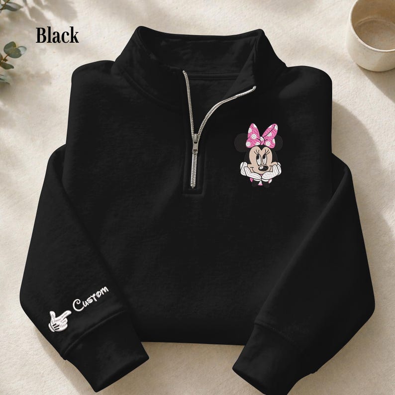Pode incluir: Camisola preta com fecho de correr de um quarto de gola. Apresenta um gr&aacute;fico da Minnie Mouse com um la&ccedil;o rosa de bolinhas. A manga esquerda tem um gr&aacute;fico branco de uma m&atilde;o e a palavra "Custom". A palavra "Black" est&aacute; no canto superior esquerdo.