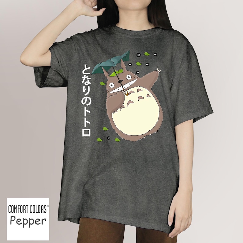 May include: Dark grey t-shirt with a cartoon Totoro character holding an umbrella. The Japanese text "となりのトトロ" is printed vertically on the left. The shirt is a Comfort Colors Pepper tee.