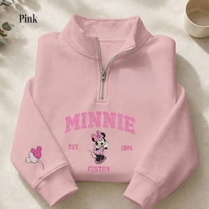 May include: A light pink quarter-zip sweatshirt featuring an embroidered Minnie Mouse graphic, the text MINNIE EST. 1928 CUSTOM, and a small balloon icon on the sleeve. The garment has a silver-toned metal zipper at the high collar neckline.