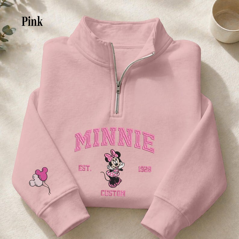 May include: A light pink quarter-zip sweatshirt featuring an embroidered Minnie Mouse graphic, the text MINNIE EST. 1928 CUSTOM, and a small balloon icon on the sleeve. The garment has a silver-toned metal zipper at the high collar neckline.