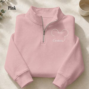 May include: Pink pullover sweatshirt with a quarter-zip neckline. The sweatshirt features a white outline of a mouse head and the word "Custom" in script. The word "Pink" is in the upper left corner.