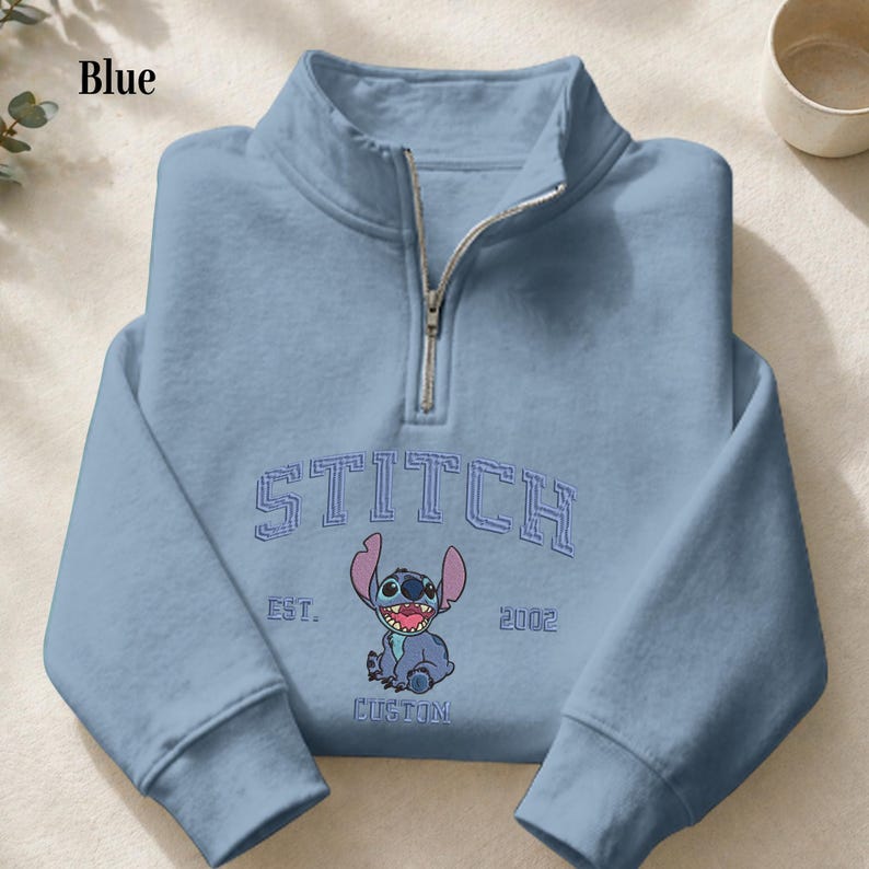 Pu&ograve; includere: Felpa blu chiaro con collo a zip a un quarto. La parte anteriore presenta la scritta "STITCH" sopra una grafica di un personaggio dei cartoni animati, con "EST. 2002" e "CUSTOM" sotto. La parola "Blue" &egrave; nell'angolo in alto a sinistra.