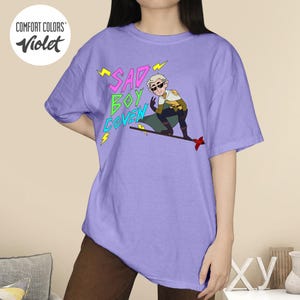 Puede incluir: Camiseta violeta Comfort Colors con el texto "SAD BOY COVEN" en rosa y verde, con detalles de rayos amarillos. La camiseta tambi&eacute;n muestra un personaje de dibujos animados.