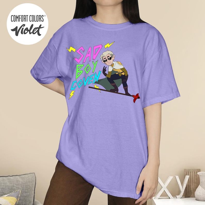 Puede incluir: Camiseta violeta Comfort Colors con el texto "SAD BOY COVEN" en rosa y verde, con detalles de rayos amarillos. La camiseta tambi&eacute;n muestra un personaje de dibujos animados.