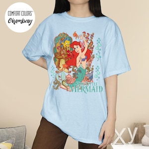 May include: Light blue t-shirt featuring a colorful graphic of Ariel from The Little Mermaid. The design includes Ariel, Flounder, and floral elements. The shirt has the text "The Little Mermaid" in a teal color.
