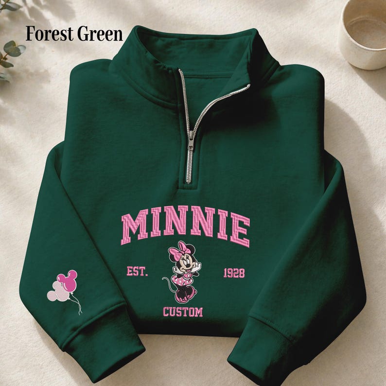 May include: A forest green quarter-zip sweatshirt featuring a pink graphic of Minnie Mouse, the text MINNIE, EST. 1928, and CUSTOM. The left sleeve displays three small pink and white balloons shaped like mouse ears. The zipper is silver-toned.