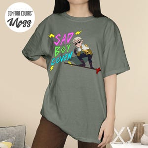 Puede incluir: Camiseta Comfort Colors verde musgo con el texto "SAD BOY COVEN" en rosa y verde, y una ilustraci&oacute;n de personaje de dibujos animados. El personaje lleva gafas de sol y un atuendo amarillo y blanco.