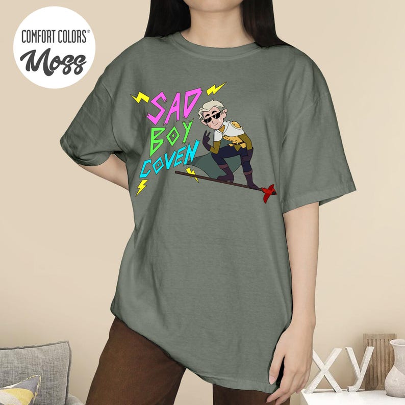 Puede incluir: Camiseta Comfort Colors verde musgo con el texto "SAD BOY COVEN" en rosa y verde, y una ilustraci&oacute;n de personaje de dibujos animados. El personaje lleva gafas de sol y un atuendo amarillo y blanco.