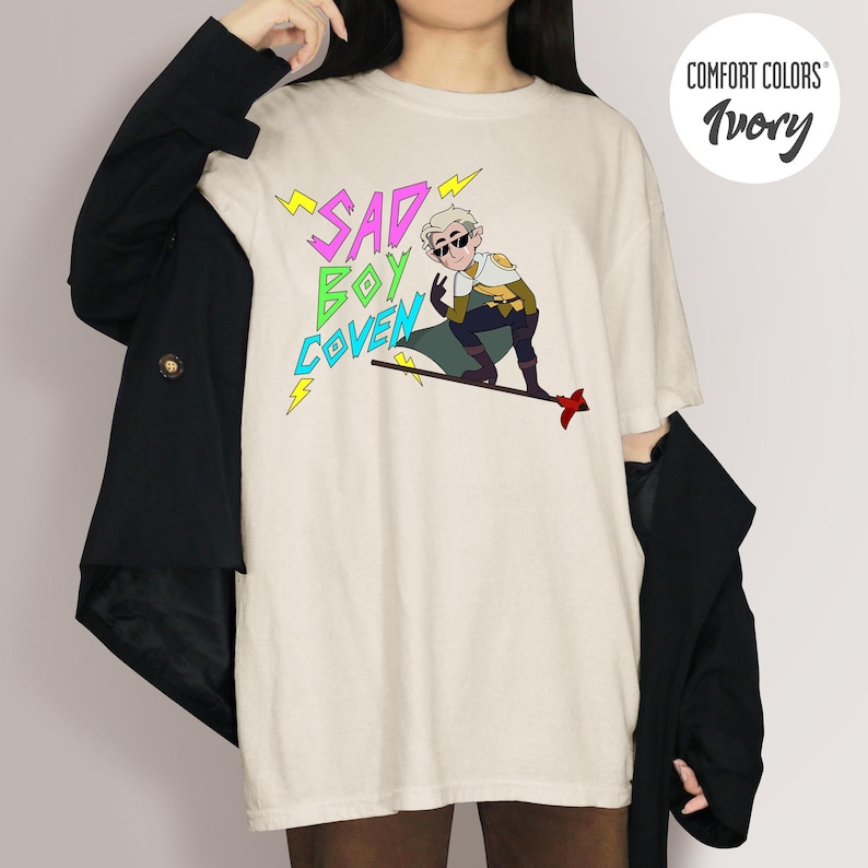 Puede incluir: Camiseta marfil Comfort Colors con un personaje de dibujos animados con gafas de sol y el texto "SAD BOY COVEN" en amarillo y verde. El personaje est&aacute; en un monopat&iacute;n con una flecha roja. Parte de una colecci&oacute;n de ropa.