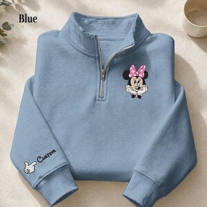 Pode incluir: Camisola azul clara com gola com fecho de correr. A camisola apresenta um design bordado da Minnie Mouse com um la&ccedil;o rosa no peito. A manga esquerda tem um gr&aacute;fico de luva branca e a palavra "Custom". A palavra "Blue" est&aacute; no canto superior esquerdo.