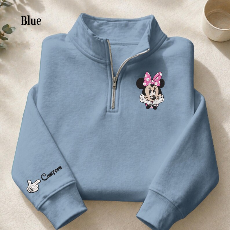 Pode incluir: Camisola azul clara com gola com fecho de correr. A camisola apresenta um design bordado da Minnie Mouse com um la&ccedil;o rosa no peito. A manga esquerda tem um gr&aacute;fico de luva branca e a palavra "Custom". A palavra "Blue" est&aacute; no canto superior esquerdo.