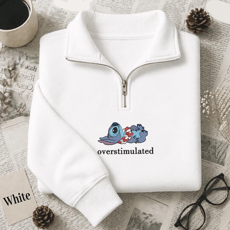 May include: A white quarter-zip sweatshirt featuring a graphic of a blue alien character wearing a red and white floral lei, lying down with the text "overstimulated" printed in black underneath the character on the center of the chest.