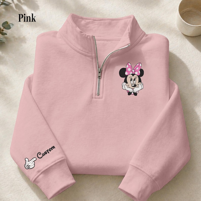 Pode incluir: Camisola rosa claro com fecho de correr de um quarto de gola, com um desenho da Minnie Mouse e um la&ccedil;o rosa com bolinhas. A manga esquerda tem um desenho personalizado com uma m&atilde;o a apontar e a palavra "Custom".