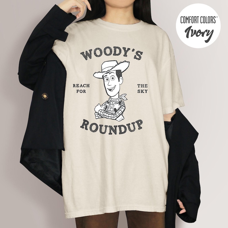 Pu&ograve; includere: T-shirt color avorio con grafica del cartone animato Woody e la scritta "WOODY'S ROUNDUP REACH FOR THE SKY". La maglietta &egrave; di Comfort Colors.