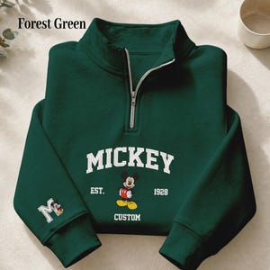 May include: A forest green quarter-zip sweatshirt featuring a white embroidered graphic of Mickey Mouse with the text MICKEY EST. 1928 CUSTOM. The right sleeve includes a white letter M with a small Mickey Mouse character icon embroidered on it.