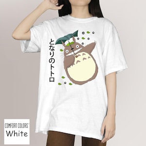 May include: White t-shirt with a cartoon Totoro character under an umbrella, with Japanese text and green leaf details. The shirt is labelled "Comfort Colors" and is a white, short-sleeved tee.